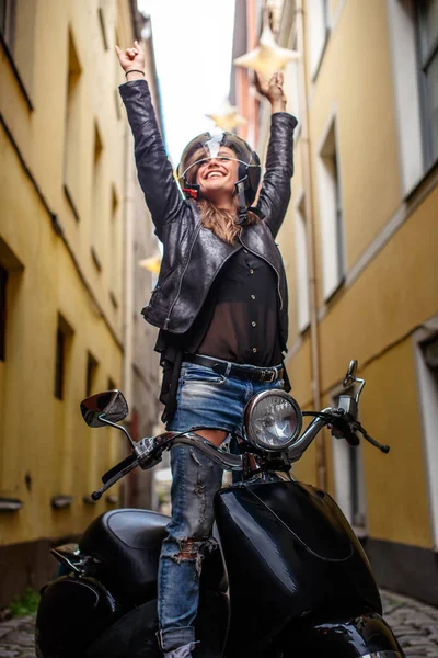 Vrolijke hipster meisje het dragen van een leren jas en gescheurde jeans permanent in een zwarte klassieke scooter op de oude smalle straat van Europa. — Stockfoto