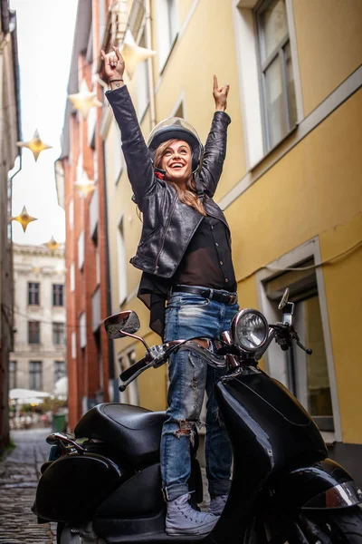 Fröhliches Hipstermädchen in Lederjacke und zerrissenen Jeans in einem schwarzen Oldtimer-Roller auf der alten engen Straße von Europa. — Stockfoto