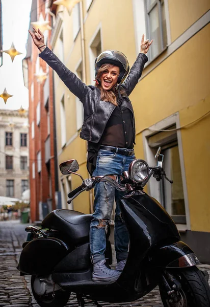Menina hipster alegre vestindo uma jaqueta de couro e jeans rasgado de pé em uma scooter clássico preto na antiga rua estreita da Europa . — Fotografia de Stock