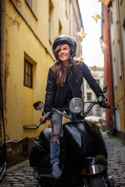 Chica hipster alegre con una chaqueta de cuero y vaqueros rasgados de pie en un scooter clásico negro en la antigua calle estrecha de Europa . — Foto de Stock