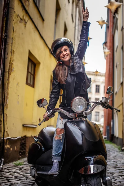 Cheerful hipster girl wearing a leather jacket and ripped jeans standing in a black classic scooter on the old narrow street of Europe. — Stock Photo, Image