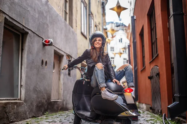 Mooi meisje het dragen van een leren jas en gescheurde jeans zittend op een zwarte klassieke scooter op de oude smalle straat van Europa. — Stockfoto