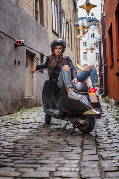 Krásná dívka, která nosí koženou bundu a roztrhané džíny sedí na černé klasické koloběžce na úzké ulici Evropy. — Stock fotografie