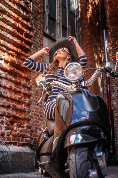 Fashionable woman wearing dress and hat sitting on a black classic Italian scooter on an old street in Europe. — Stock Photo, Image