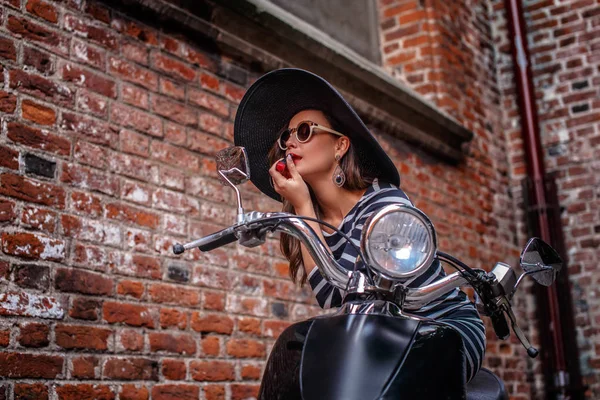 Donna alla moda indossa abito e cappello seduto accanto a uno scooter nero e dipinge le labbra con rossetto guardando nello specchio retrovisore . — Foto Stock