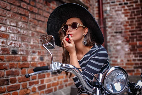Modische Frau mit Kleid und Hut sitzt stehend neben einem schwarzen Roller und bemalt ihre Lippen mit Lippenstift, schaut in den Rückspiegel. — Stockfoto