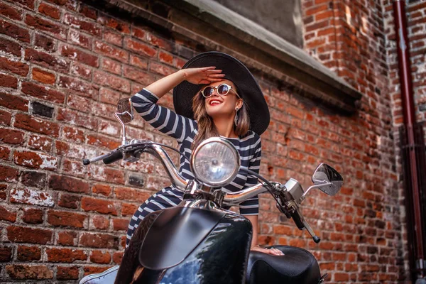 Mulher alegre vestindo roupas de moda apoiando-se em uma scooter italiana clássico preto e olhando para longe em uma rua velha na Europa . — Fotografia de Stock