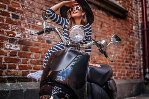 Wanita ceria yang mengenakan pakaian mode bersandar pada skuter Italia klasik hitam dan memandang jauh di sebuah jalan tua di Eropa . — Stok Foto
