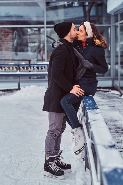 Junges attraktives verliebtes Paar, Date auf der Eisbahn, ein Mädchen sitzt auf einer Leitplanke und küsst ihren Freund. — Stockfoto