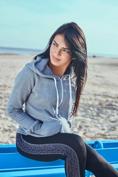 Una bella ragazza con la pelle abbronzata indossa una felpa grigia con cappuccio che distoglie lo sguardo e si tiene le mani in tasca mentre si siede su una panchina in spiaggia — Foto Stock