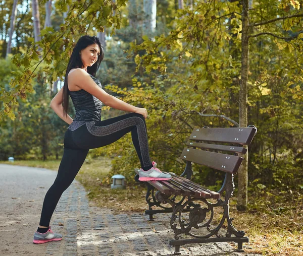 在秋季公园户外跑步前, 年轻的健身女子穿着运动服热身, 伸展双腿 — 图库照片
