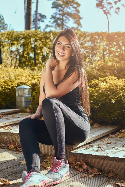 Un'affascinante ragazza fitness con la pelle abbronzata che indossa abbigliamento sportivo seduta su gradini di legno al parco, sorridente e guardando una macchina fotografica — Foto Stock