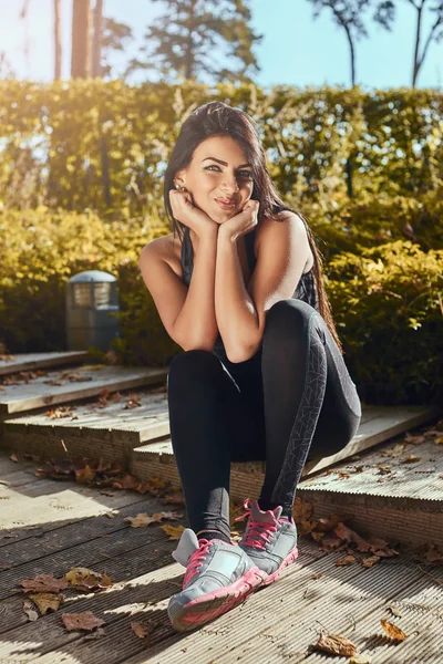 Un'affascinante ragazza fitness con la pelle abbronzata che indossa abbigliamento sportivo seduta su gradini di legno al parco, sorridente e guardando una macchina fotografica — Foto Stock