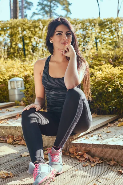 Una hermosa chica con piel bronceada usando ropa deportiva sentada en escalones de madera en el parque — Foto de Stock