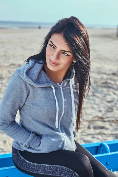 Tabaklanmış deri bir gri kapşonlu uzağa arıyorsunuz ve tutan elleri ceplerinde plajda bir bankta otururken giyen güzel bir kızla — Stok fotoğraf