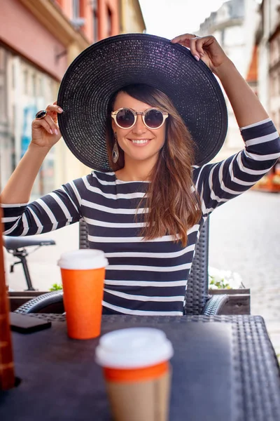 Retrato de uma jovem mulher na moda usando óculos de sol e chapéu elegante sentado em um café de rua de verão . — Fotografia de Stock