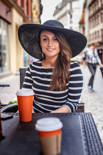 Ritratto di una donna sorridente che indossa un vestito e un cappello alla moda seduta in un caffè di strada estivo . — Foto Stock