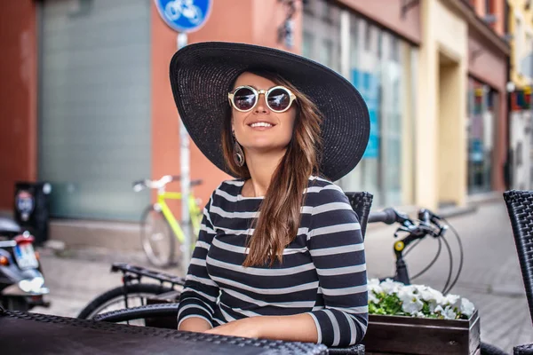 Elegantemente vestido jovem mulher vestindo um chapéu elegante e óculos de sol sentado em um café de rua de verão . — Fotografia de Stock