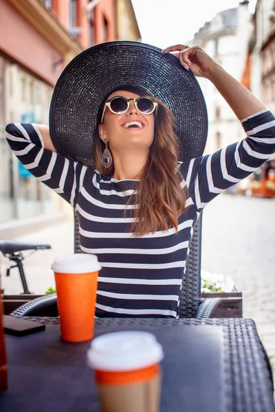 Retrato de uma jovem mulher na moda usando óculos de sol e chapéu elegante sentado em um café de rua de verão . — Fotografia de Stock