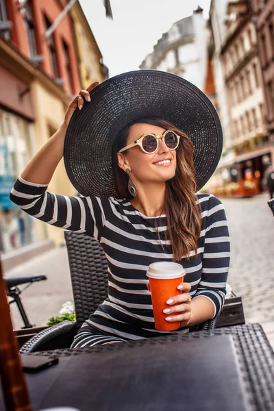 Ritratto di una giovane donna alla moda che indossa occhiali da sole e cappello elegante seduta in un caffè di strada estivo . — Foto Stock