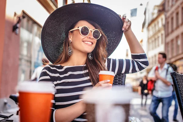 Portret van een jonge modieuze vrouw dragen zonnebril en stijlvolle hoed zitten in een zomer straat Cafe. — Stockfoto