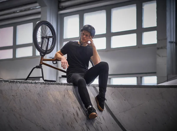 Bmx гонщик розслабляється після тренування трюків з велосипедом в скейтпарку в приміщенні — стокове фото