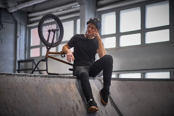 Bmx jinete relajante después de practicar trucos con su bicicleta en un skatepark en el interior — Foto de Stock