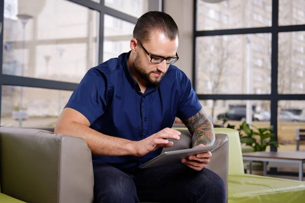 Hombre de negocios barbudo guapo vestido con una camisa azul sentado en el sofá en la oficina, trabajando con la tableta digital —  Fotos de Stock