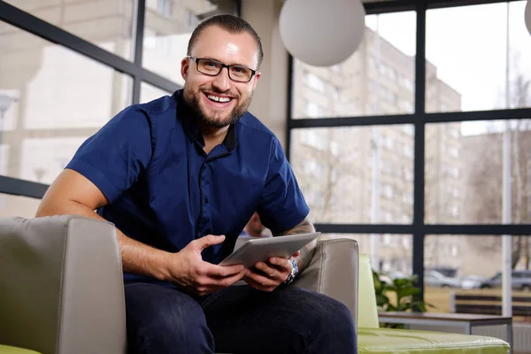 Homme d'affaires prospère vêtu d'une chemise bleue assis sur le canapé dans le bureau, tenant une tablette, souriant et regardant une caméra — Photo