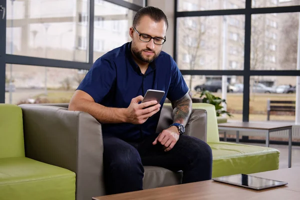 Empresario barbudo enfocado vestido con una camisa azul sentado en el sofá de la oficina, usando el teléfono inteligente — Foto de Stock