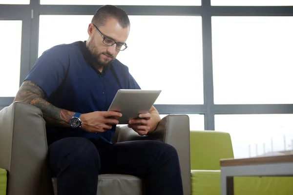 Pohledný podnikatel vousatý oblečený v modré košili, sedící na gauči v kanceláři, práce s digitálním tabletu — Stock fotografie