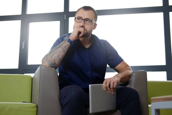 Schöner bärtiger Geschäftsmann in blauem Hemd sitzt auf der Couch im Büro und hält das digitale Tablet in der Hand — Stockfoto