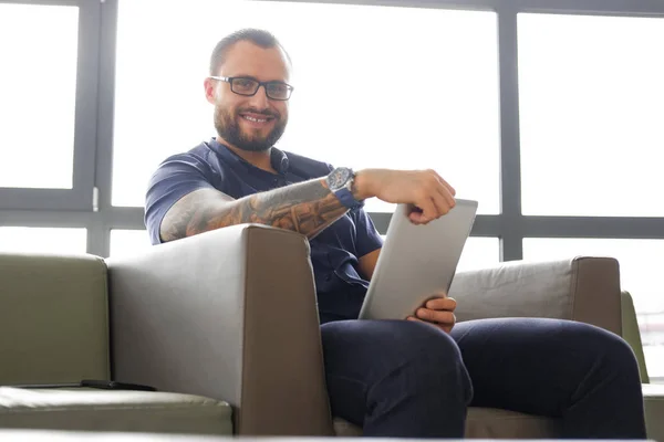 Homme d'affaires prospère vêtu d'une chemise bleue assis sur le canapé dans le bureau, tenant une tablette, souriant et regardant une caméra — Photo