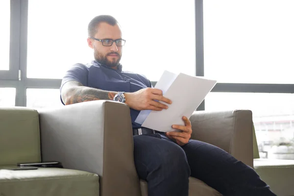 Homme d'affaires barbu concentré vêtu d'une chemise bleue assis sur le canapé et examine les documents importants dans le bureau — Photo