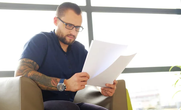 Homme d'affaires barbu concentré vêtu d'une chemise bleue assis sur le canapé et examine les documents importants dans le bureau — Photo