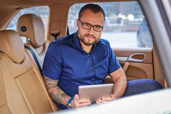 Homem de negócios barbudo sentado em um banco de trás no carro de luxo, segurando um tablet e olhando para uma câmera — Fotografia de Stock