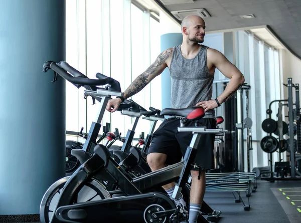 Портрет лысого спортсмена с татуировкой на руке, стоящего рядом с велотренажером в современном фитнес-клубе — стоковое фото