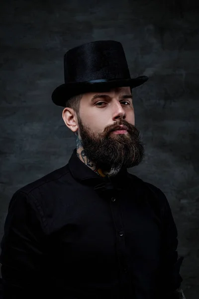 Retrato de un hombre barbudo anticuado con tatuajes en el cuello con un sombrero de copa, aislado sobre un fondo oscuro. Captura de estudio —  Fotos de Stock