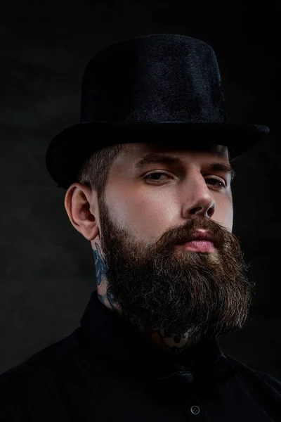 Retrato de close-up de um homem barbudo à moda antiga com tatuagens no pescoço usando um chapéu superior, isolado em um fundo escuro. Estúdio — Fotografia de Stock