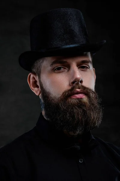 Retrato de close-up de um homem barbudo à moda antiga com tatuagens no pescoço usando um chapéu superior, isolado em um fundo escuro. Estúdio — Fotografia de Stock