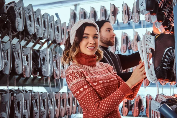 Young couple wearing warm clothes standing near rack with many pairs of skates, choosing his size