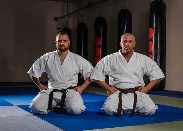 Two karate fighters in kimono sitting on their knees ready to start fighting in the gym — Stock Photo, Image