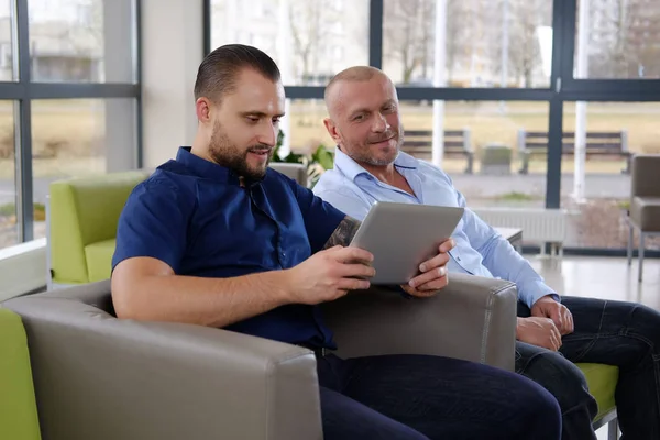 Cheerful coworkers are watching something funny in the tablet sitting on a couch in the office — Stock Photo, Image