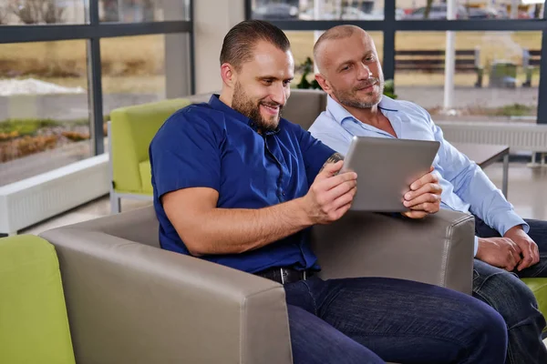 Compañeros alegres están viendo algo divertido en la tableta sentados en un sofá en la oficina —  Fotos de Stock