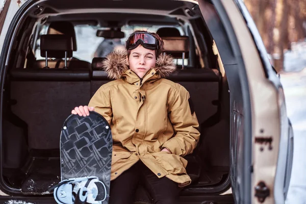 Een tiener snowboarder gekleed in warme kleding, zittend op de kofferbak van een auto — Stockfoto