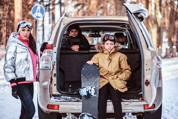 Una hermosa mujer y sus hijos vestidos con ropa de abrigo sentados en el maletero de un coche — Foto de Stock