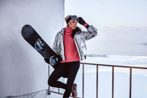 Uma bela mulher vestindo roupas quentes e óculos posando com snowboard, olhando para longe enquanto se inclina em um corrimão perto de uma praia nevada — Fotografia de Stock