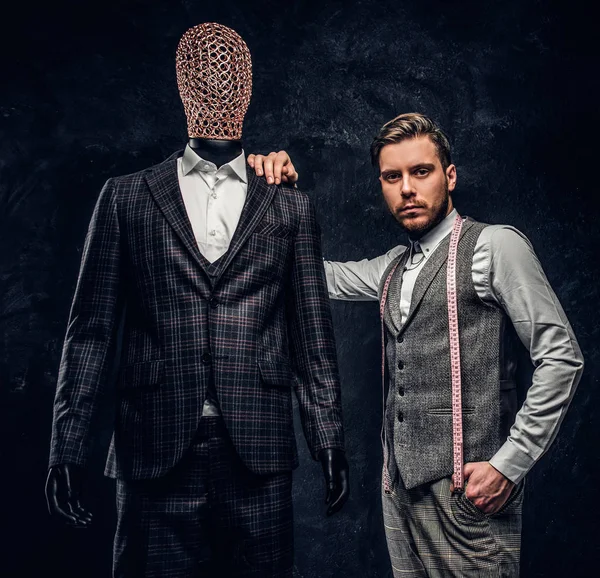 Un joven diseñador de moda confiado posando junto a un maniquí en traje exclusivo hecho a medida para hombre en un estudio de sastre oscuro — Foto de Stock