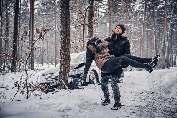 Cara das últimas forças carrega sua namorada ferida em um acidente em uma estrada nevada na floresta — Fotografia de Stock