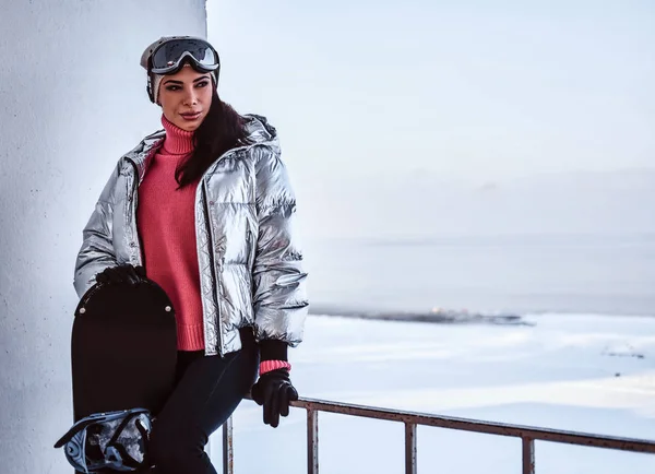 Uma bela mulher vestindo roupas quentes e óculos posando com snowboard, olhando para longe enquanto se inclina em um corrimão perto de uma praia nevada — Fotografia de Stock